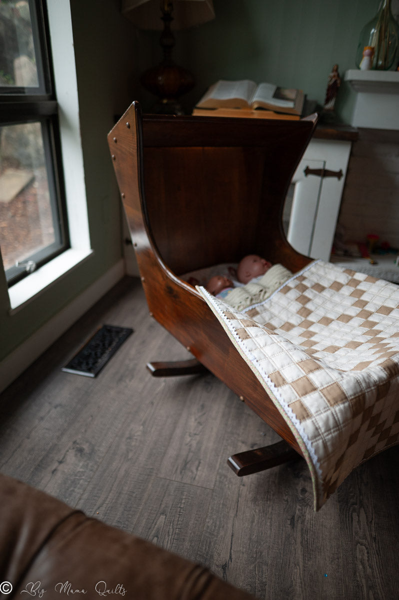 Doll sleeping in a wooden rocking cradle with a quilt draped over it, in a room with a window.